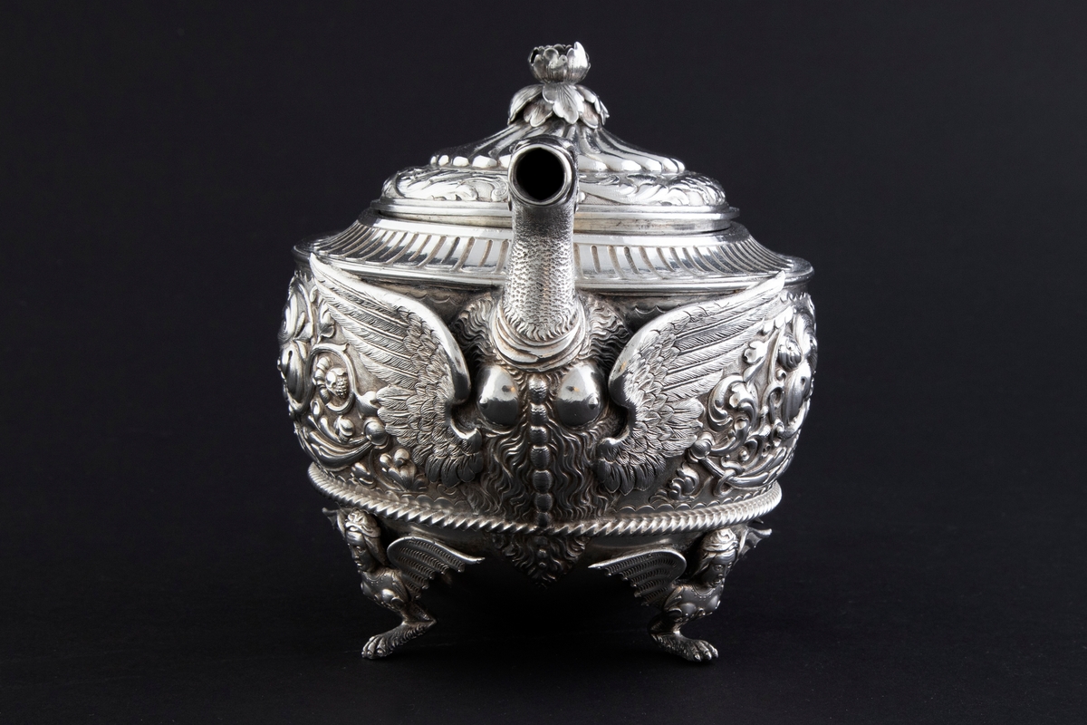 Tekanne i sølv. Tilhærmet halvkuleformet korpus, dekorert med rankeornamenter i relieff. Opprinnelig har overflaten mellom rankene vært oksidert. Kannen bæres av fire små ben, utformet som harpyier-lignede vesener, hvor vingene utgjør ansatsen til korpus. Svungen hank med plastisk utformet bladornament, samt tut med en griffelignende utforming. Sirkulært hengslet lokk med konsentrisk plastisk dekor og blomsterformet lokknapp.