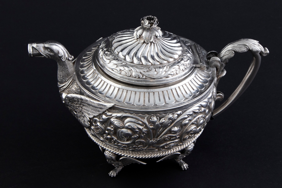 Tekanne i sølv. Tilhærmet halvkuleformet korpus, dekorert med rankeornamenter i relieff. Opprinnelig har overflaten mellom rankene vært oksidert. Kannen bæres av fire små ben, utformet som harpyier-lignede vesener, hvor vingene utgjør ansatsen til korpus. Svungen hank med plastisk utformet bladornament, samt tut med en griffelignende utforming. Sirkulært hengslet lokk med konsentrisk plastisk dekor og blomsterformet lokknapp.