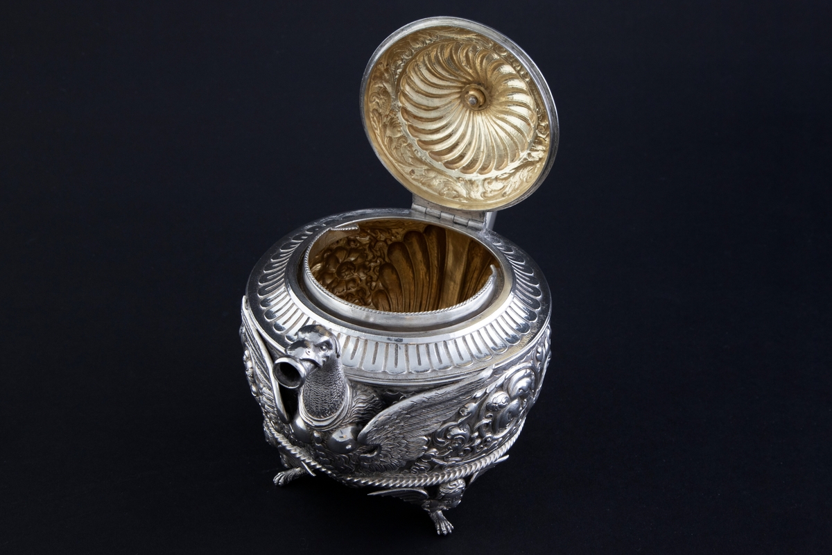 Tekanne i sølv. Tilhærmet halvkuleformet korpus, dekorert med rankeornamenter i relieff. Opprinnelig har overflaten mellom rankene vært oksidert. Kannen bæres av fire små ben, utformet som harpyier-lignede vesener, hvor vingene utgjør ansatsen til korpus. Svungen hank med plastisk utformet bladornament, samt tut med en griffelignende utforming. Sirkulært hengslet lokk med konsentrisk plastisk dekor og blomsterformet lokknapp.