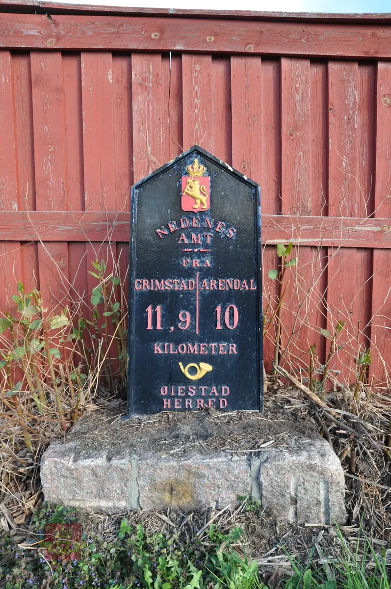 Milepæl i støpejern ved riksveg 407 hovedparsell 02 km 3,158. 
Motiv: Riksvåpen med løve og krone + posthorn. Tekst: NEDENES AMT - FRA GRIMSTAD 11,9 - ARENDAL 10 KILOMETER - ØIESTAD HERRED. Datering ukjent. 
Registrert for Statens vegvesen av Næs Jernverksmuseum i 2012 som del av prosjekt med registrering av milepeler og grensemerker mm. 

Stedsnavn: Væding