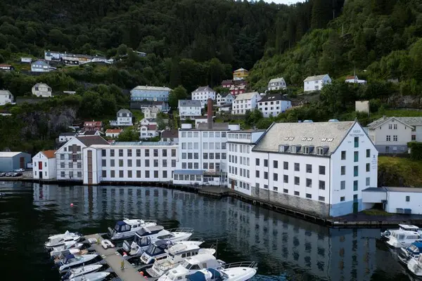 Hvite ruvende fabrikklokaler i flere etasjer liggende helt tett ved sjøen med bratt li bak