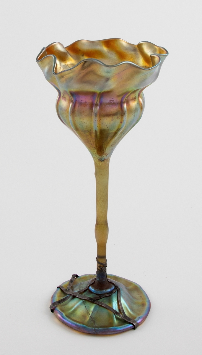 Høyreist vase i farvet glass, hvis form trolig alluderer til en blomst. Både kupa og fot er kraftig irisert, hvor det oppstår skimmrende gullfargede partier med innslag av lilla, blått og grønt. Konisk kupa med organisk utforming, samt kraftig bølgende formet munningsrand. Vertikale riller på nedre del av kupa, som bæres av en tynn stett. Sirkulær fotplate, noe hevet på midten. Fotplaten har et tverrgående brudd, som senere er limt, og  er holdt sammen av metallbånd.