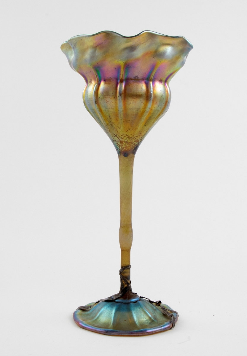 Høyreist vase i farvet glass, hvis form trolig alluderer til en blomst. Både kupa og fot er kraftig irisert, hvor det oppstår skimmrende gullfargede partier med innslag av lilla, blått og grønt. Konisk kupa med organisk utforming, samt kraftig bølgende formet munningsrand. Vertikale riller på nedre del av kupa, som bæres av en tynn stett. Sirkulær fotplate, noe hevet på midten. Fotplaten har et tverrgående brudd, som senere er limt, og  er holdt sammen av metallbånd.