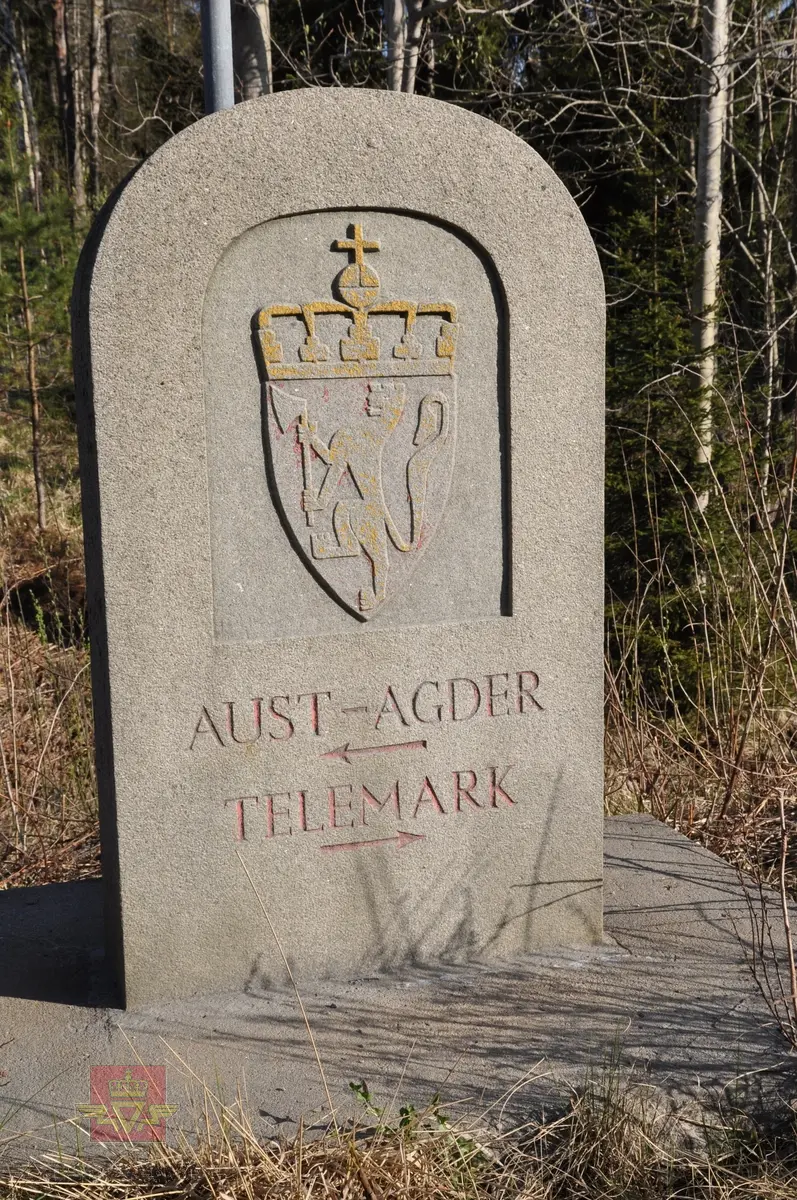 Grensestein fylkesgrensen mellom Aust-Agder og Telemark. 
Motiv: Riksvåpen i relieff + tekst:  AUST-AGDER - TELEMARK. 
Registrert for Statens vegvesen av Næs Jernverksmuseum i 2012 som del av prosjekt med registrering av milepeler og grensemerker mm. 

Stedsnavn: Østerholtheia