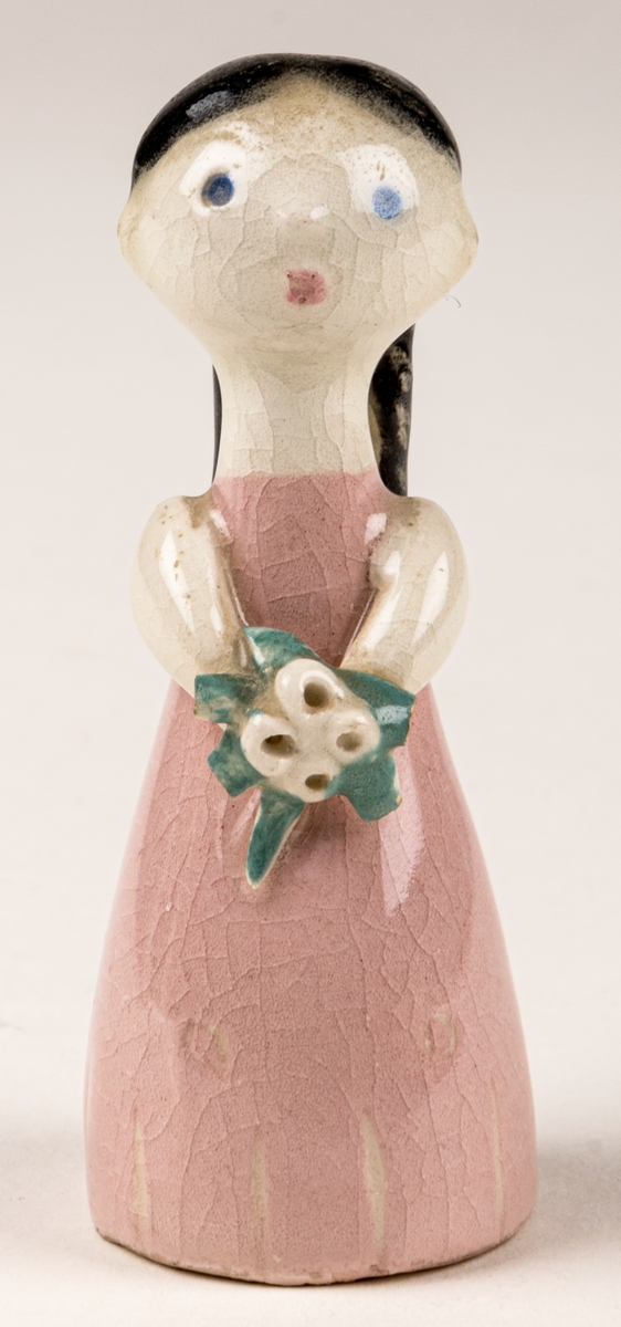 Brudtärna till figuringrupp Brudfölje. Tärnan har mörkt hår och rosa klänning. Formgiven av Dorothy Clough 1955, Gefle Porslinsfabrik. På undersidan svaga rester av stämpeltryckt logga med GEFLE och tre rundugnar.