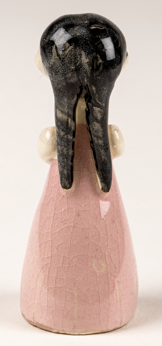 Brudtärna till figuringrupp Brudfölje. Tärnan har mörkt hår och rosa klänning. Formgiven av Dorothy Clough 1955, Gefle Porslinsfabrik. På undersidan svaga rester av stämpeltryckt logga med GEFLE och tre rundugnar.