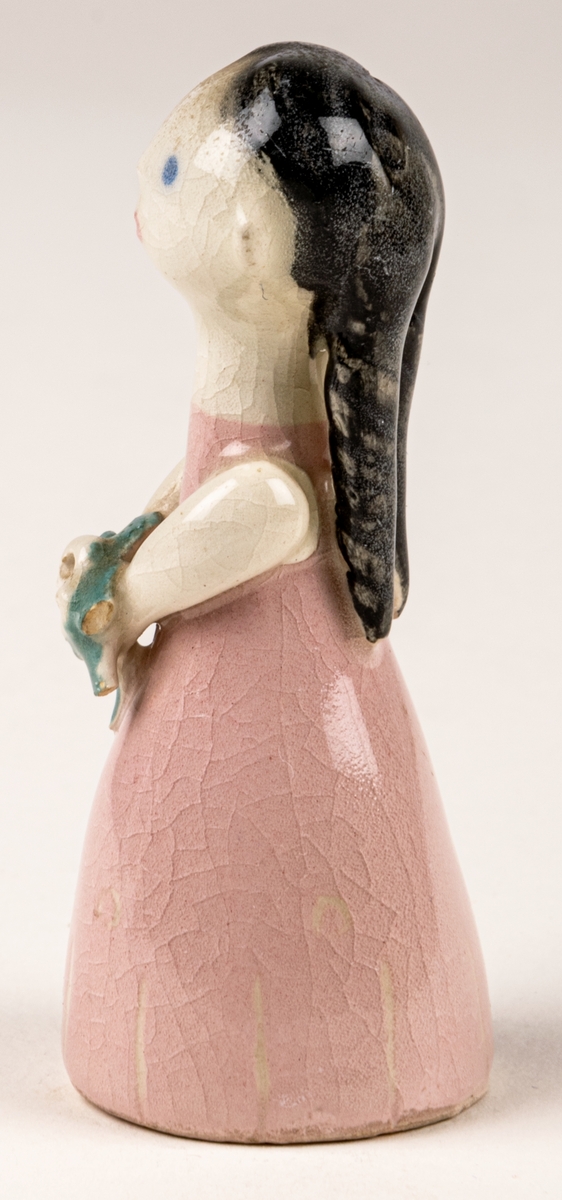 Brudtärna till figuringrupp Brudfölje. Tärnan har mörkt hår och rosa klänning. Formgiven av Dorothy Clough 1955, Gefle Porslinsfabrik. På undersidan svaga rester av stämpeltryckt logga med GEFLE och tre rundugnar.