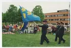 Fyra militärer förflyttar sig med två blå gula fanor.
Publik och länsstyrelsens byggnad finns i bakgrunden.
