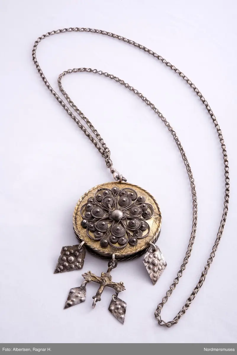 Lavendeldåse med gitterverkblomst på lokket og feste for lenka. Fra dåsen henger løv og et krusifiks med løv. Kjeden har karabinkrok.