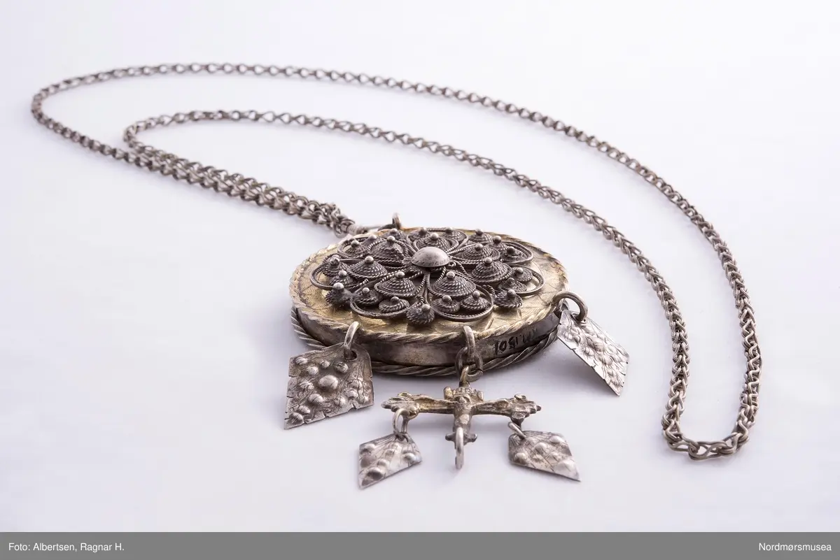 Lavendeldåse med gitterverkblomst på lokket og feste for lenka. Fra dåsen henger løv og et krusifiks med løv. Kjeden har karabinkrok.