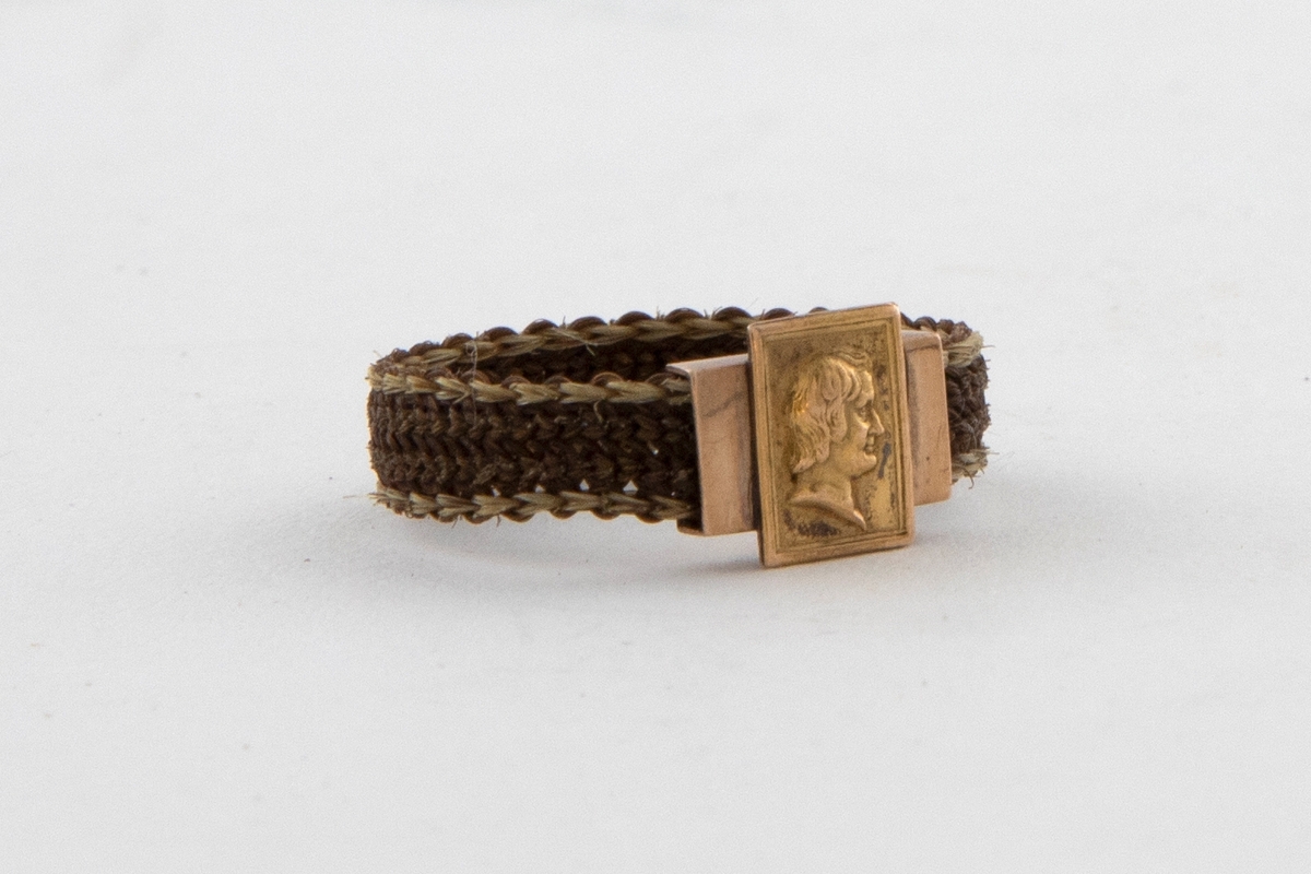 Fingerring flettet av hår. Jevnt bredt bånd som er tett flettet i lyst brunt hår med en smal kant av blondt hår på hver side. Ringen har et rektangulært gullbeslag på midten. En gullplate med et profilhode av en mann er festet tvers over beslaget.