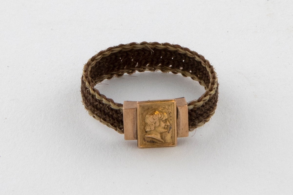 Fingerring flettet av hår. Jevnt bredt bånd som er tett flettet i lyst brunt hår med en smal kant av blondt hår på hver side. Ringen har et rektangulært gullbeslag på midten. En gullplate med et profilhode av en mann er festet tvers over beslaget.