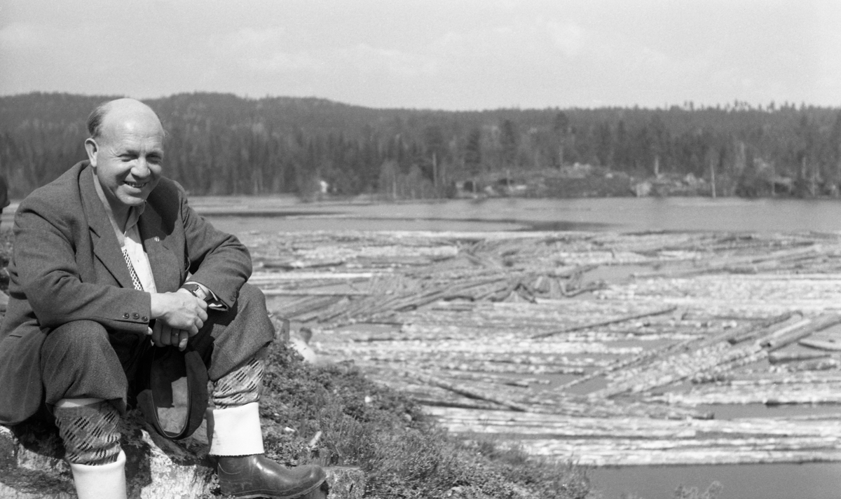 Gardbruker Arne Holth (1913-1995) fra Gardvik i Nord-Odal i Hedmark, fotografert sittende på en bakkekam ved Gåsvatnet i Nord-Odal. På vannspeilet lå det en betydelig mengde tømmer. Holth var kledd i dressjakke og nikkersbukser.