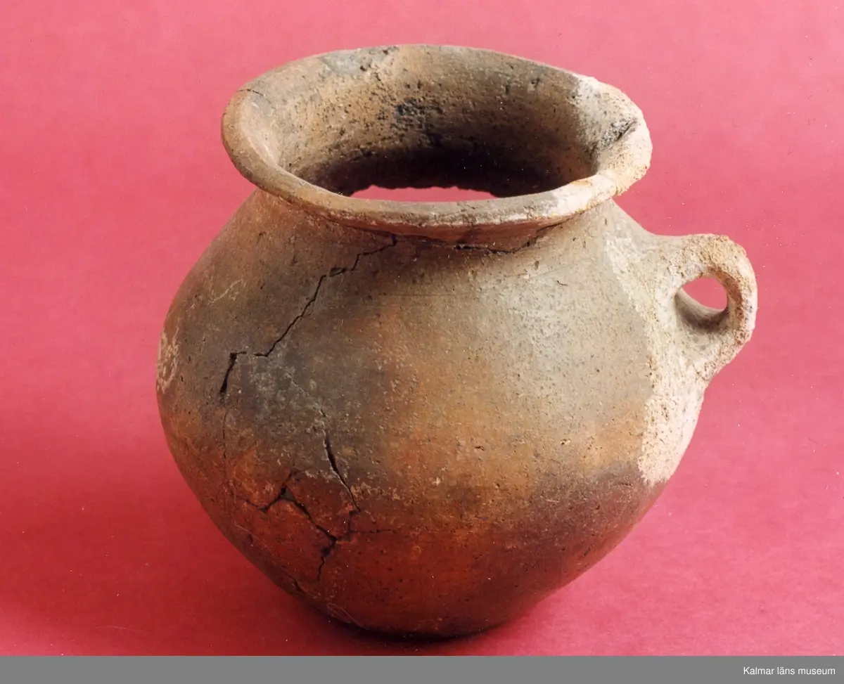 KLM 2308. Gravurna. Rundbukigt lerkärl med utsvängd mynningskant, ett öra. Ornamentik i form av gropar och oregelbundna linjer under mynningsranden. Skadad. Ihoplimmad.