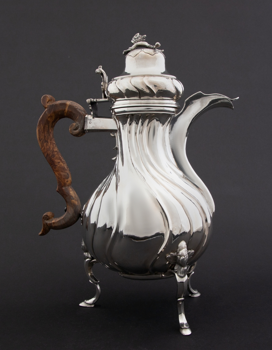 Pæreformet kaffekanne i sølv, som hviler på tre høye svungne bein, hvor ansatsen til korpus er markert med en støpt blomst. Korpus dekorert med sveifede knekk, som fortsetter på nedre del av det hjelmformede lokket. Øvre del  av lokket er udekorert. Det krones med en lokknapp som er utformet som en todelt gren, hvorpå det ligger et ulvehode med en fugl i munnen. Lokkåpneren er utformet som et tre-fliket blad, hvor to av flikene er opprullet.  Kannen har svunget hank i et mørkt treslag, som er festet til korpus med en sølvskrue.