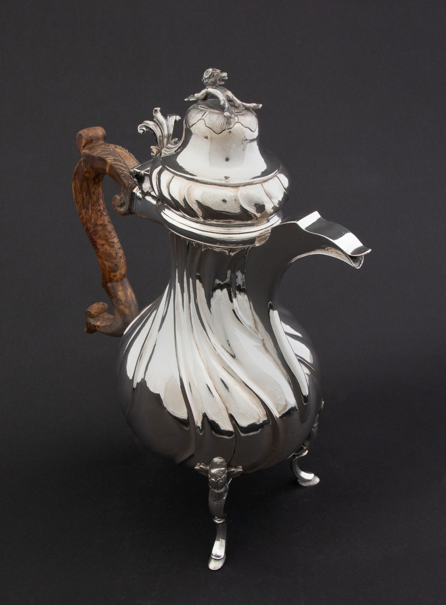 Pæreformet kaffekanne i sølv, som hviler på tre høye svungne bein, hvor ansatsen til korpus er markert med en støpt blomst. Korpus dekorert med sveifede knekk, som fortsetter på nedre del av det hjelmformede lokket. Øvre del  av lokket er udekorert. Det krones med en lokknapp som er utformet som en todelt gren, hvorpå det ligger et ulvehode med en fugl i munnen. Lokkåpneren er utformet som et tre-fliket blad, hvor to av flikene er opprullet.  Kannen har svunget hank i et mørkt treslag, som er festet til korpus med en sølvskrue.