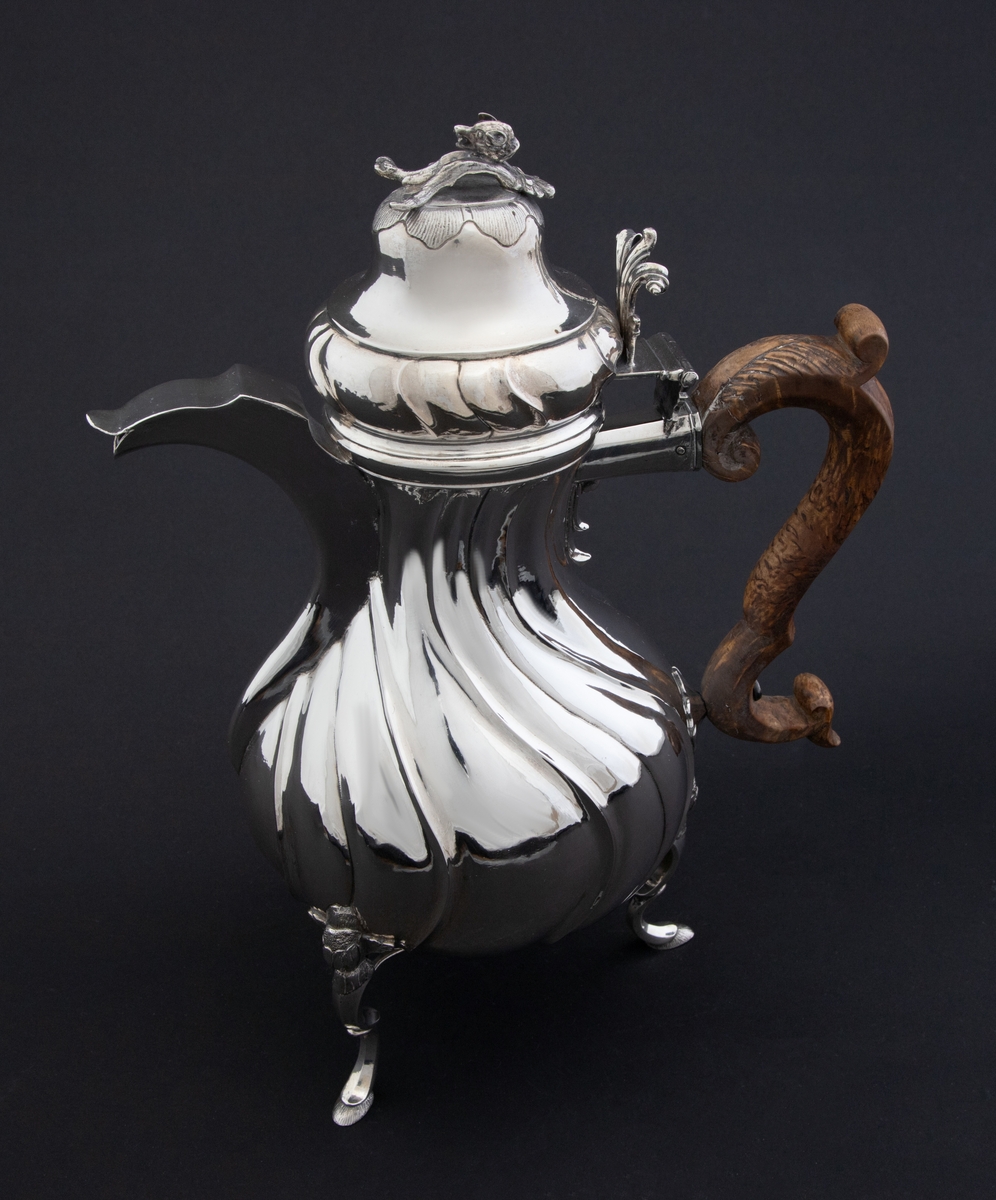 Pæreformet kaffekanne i sølv, som hviler på tre høye svungne bein, hvor ansatsen til korpus er markert med en støpt blomst. Korpus dekorert med sveifede knekk, som fortsetter på nedre del av det hjelmformede lokket. Øvre del  av lokket er udekorert. Det krones med en lokknapp som er utformet som en todelt gren, hvorpå det ligger et ulvehode med en fugl i munnen. Lokkåpneren er utformet som et tre-fliket blad, hvor to av flikene er opprullet.  Kannen har svunget hank i et mørkt treslag, som er festet til korpus med en sølvskrue.