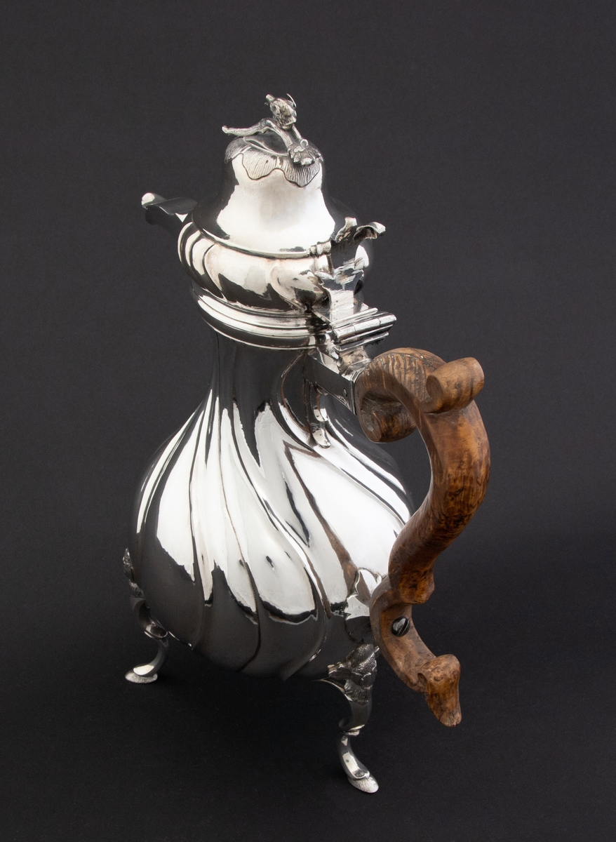 Pæreformet kaffekanne i sølv, som hviler på tre høye svungne bein, hvor ansatsen til korpus er markert med en støpt blomst. Korpus dekorert med sveifede knekk, som fortsetter på nedre del av det hjelmformede lokket. Øvre del  av lokket er udekorert. Det krones med en lokknapp som er utformet som en todelt gren, hvorpå det ligger et ulvehode med en fugl i munnen. Lokkåpneren er utformet som et tre-fliket blad, hvor to av flikene er opprullet.  Kannen har svunget hank i et mørkt treslag, som er festet til korpus med en sølvskrue.