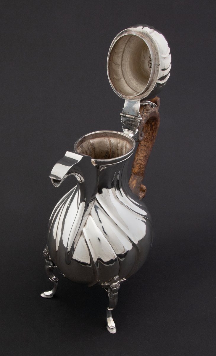 Pæreformet kaffekanne i sølv, som hviler på tre høye svungne bein, hvor ansatsen til korpus er markert med en støpt blomst. Korpus dekorert med sveifede knekk, som fortsetter på nedre del av det hjelmformede lokket. Øvre del  av lokket er udekorert. Det krones med en lokknapp som er utformet som en todelt gren, hvorpå det ligger et ulvehode med en fugl i munnen. Lokkåpneren er utformet som et tre-fliket blad, hvor to av flikene er opprullet.  Kannen har svunget hank i et mørkt treslag, som er festet til korpus med en sølvskrue.