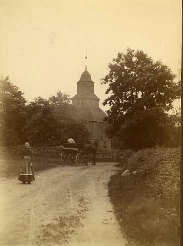 En hästskjuts är på väg mot Okome gamla kyrka och i förgrunden står en kvinna med randigt förkläde vänd mot fotografen. Okome nya kyrka uppfördes 1891.