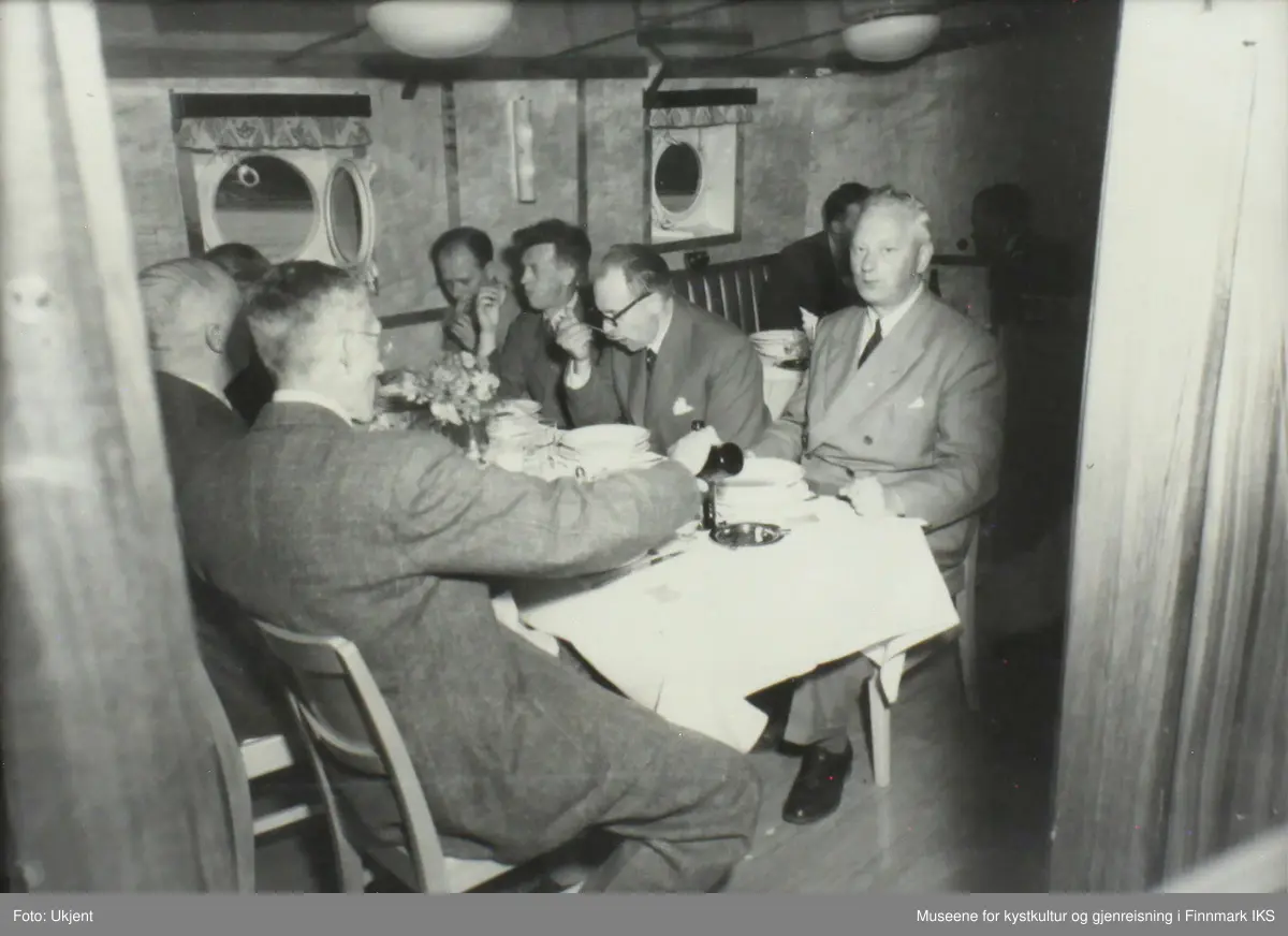 Flere menn spiser i messa til motorskipet M/S Ingøy. I anledning til avlevering fra Trosvik verksted i 1949. De er kledd i dress. En av dem kan være Peder "Pelle" Bang.
Bordet man ser er festlig dekket med hvit duk, blomster og fin porselen. 
Det er halvkuleformete lamper i taket og man ser to kuøyer, runde skipsvinduer.Veggene ser ut å være kledd med trefinér. Møblene er av tre.