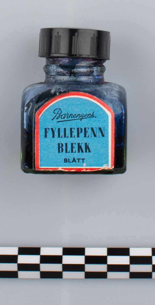 Glassflaske med tørkede rester av formentlig blått blekk. Sort skrulokk i kunstmateriale. Etikett på en side.