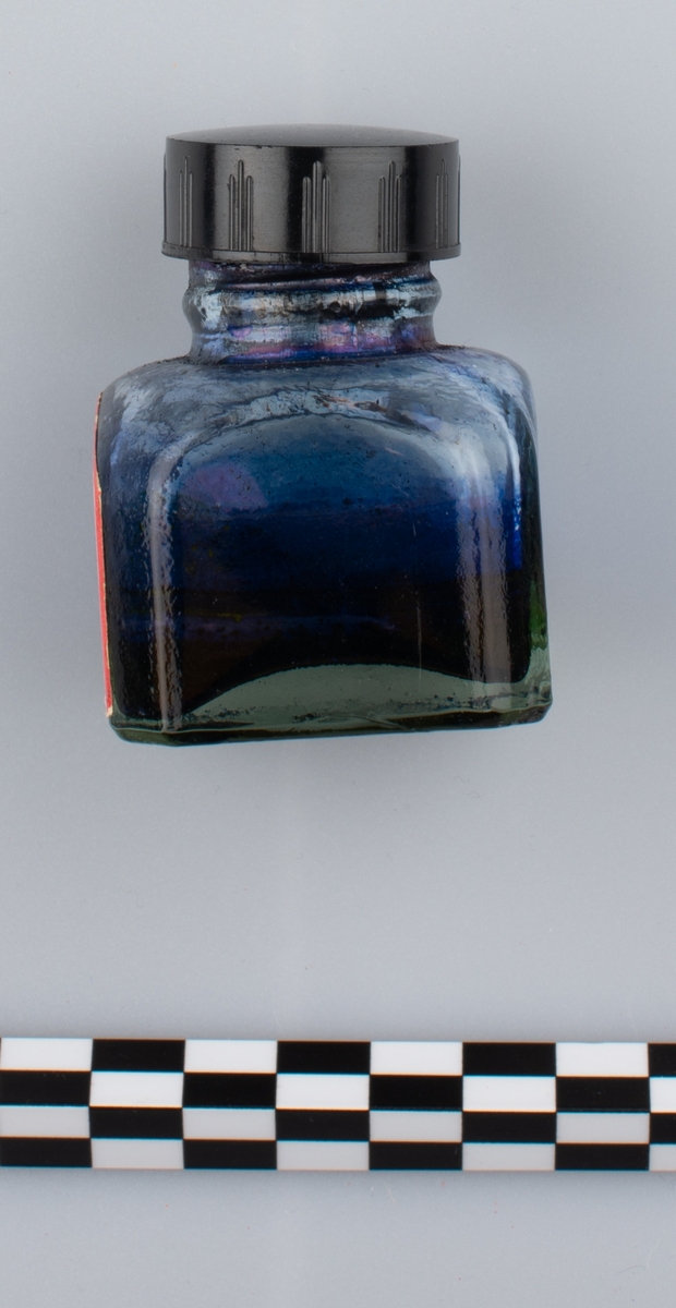 Glassflaske med tørkede rester av formentlig blått blekk. Sort skrulokk i kunstmateriale. Etikett på en side.