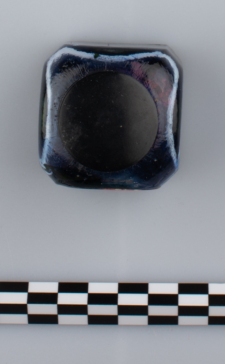 Glassflaske med tørkede rester av formentlig blått blekk. Sort skrulokk i kunstmateriale. Etikett på en side.