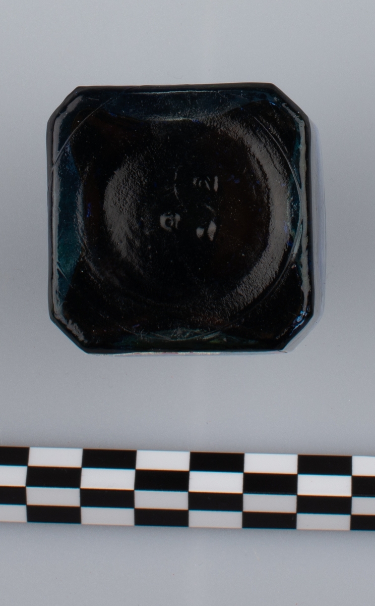 Glassflaske med tørkede rester av formentlig blått blekk. Sort skrulokk i kunstmateriale. Etikett på en side.
