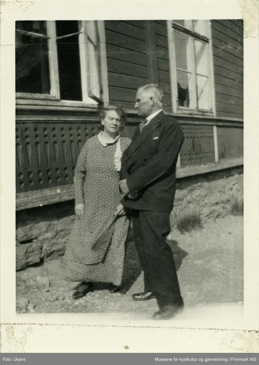 En kvinne og en mann står ved siden av en bygning.  Bygningens sokkel er pyntet med tilskjærte ornamenter, et vindu er åpent. Paret er snudd lett mot hverandre, som i en samtale. Vinden tar i hennes mønstret kjole og i håret.