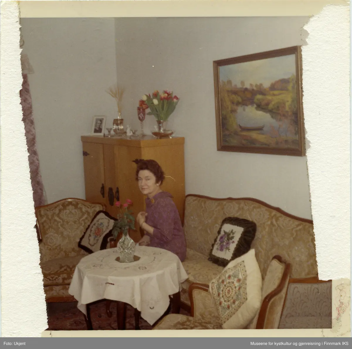 En kvinne sitter på sofaen i stua. Hun har en lilla kjole på seg og ser vennlig mot fotografen. På veggen er det et landskapsmaleri. På skapet bak henne står det et innrammet fotografi, en vase med tulipaner og annen pynt. Også det runde stuebordet er pyntet med en hvit duk og en krystallvase med tre røde roser i. Både sofaen og lenestolene er pyntet med broderte puter.