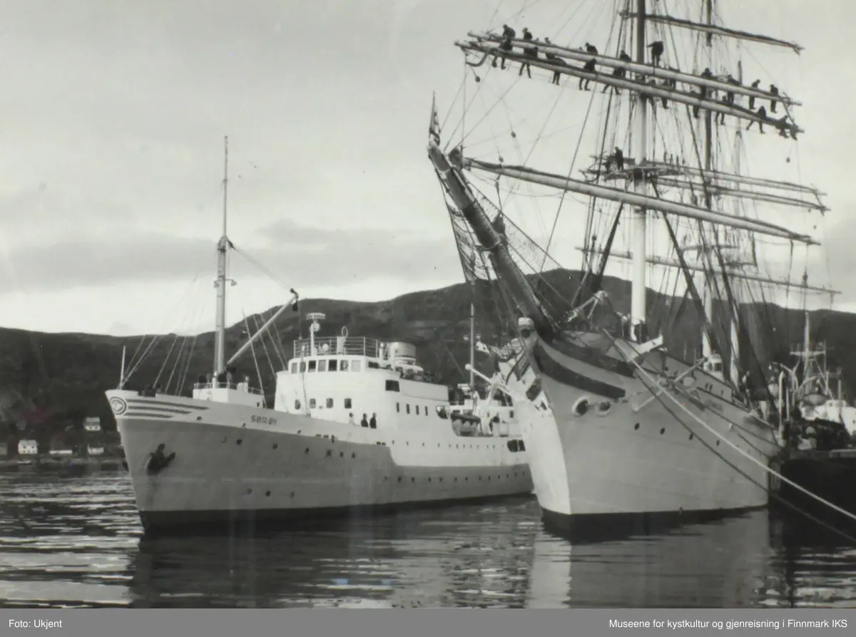 FFR - båten M/S Sørøy og tremasteren "Statsraat Lehmkuhl" ligger i havna i Hammerfest. Ombord på rutebåten står flere personer og ser på hav som foregår på den store seilbåten. Der står det mange personer på bommene.