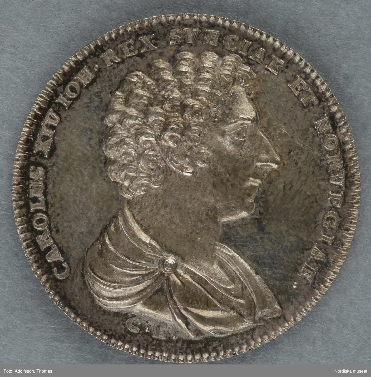 Huvudliggare:
"Minnespenningar. 3 st. I silfver. Öfver Carl XIV Johan.
a. Jfr. Hildebrand sid. 336, nr 29
b. Jfr. -"- sid.337, nr 25
c. Jfr. -"- sid 364, nr 77a"

B:
Minnespenning av silver, till minne av Carl XIV Johans kröning till kung Stockholms storkyrka den 11 maj 1818. På åtsidan kungens bröstbild och texten: "CARL XIV JOHAN SV. OCH NORR: K: KRÖNT ÅR 1818". På frånsidan kungens valspråk: "FOLKETS KÄRLEK MIN BELÖNING" inom en krans av hälften ek (symbolväxt för Norge) till hälften oliv (Sverige).
 Denna penning kastades ut bland folket på stadens torg och öppna platser vid kröningen, enligt gammalt bruk.
Carl XIV Johan var unionskung och kung över båda kungarikena Sverige och Norge.
Se Hildebrand, del 2, nr.25. Jämför inv.nr. 29 574b, som är motsvarande minnespenning från Carl XIV Johans kröning i Trondheims domkyrka samma år.
Maria Maxén/2005