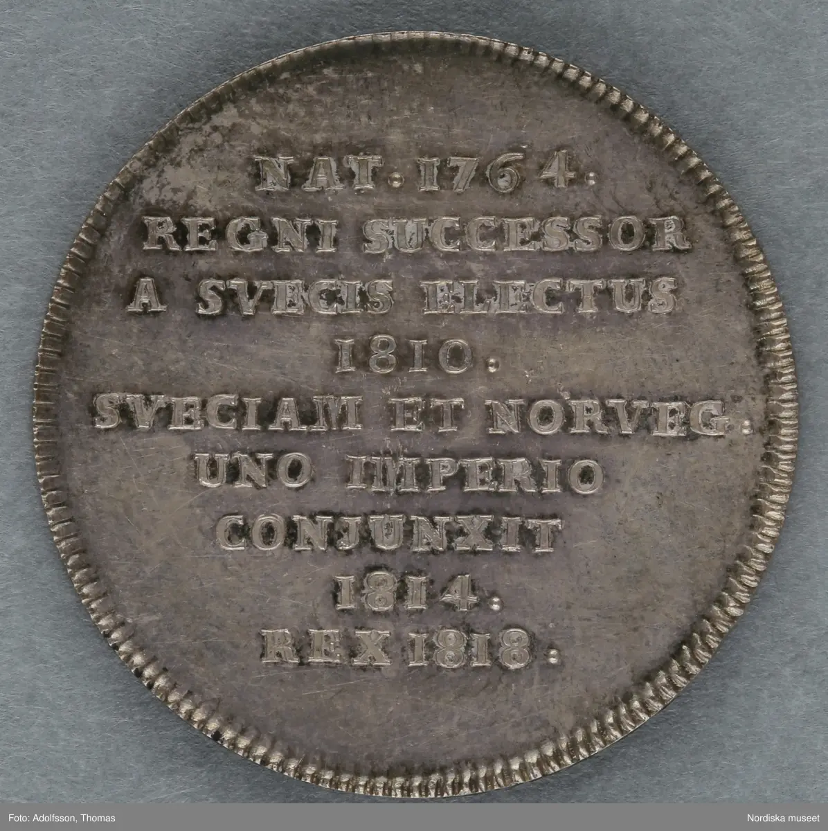 Huvudliggare:
"Minnespenningar. 3 st. I silfver. Öfver Carl XIV Johan.
a. Jfr. Hildebrand sid. 336, nr 29
b. Jfr. -"- sid.337, nr 25
c. Jfr. -"- sid 364, nr 77a"

B:
Minnespenning av silver, till minne av Carl XIV Johans kröning till kung Stockholms storkyrka den 11 maj 1818. På åtsidan kungens bröstbild och texten: "CARL XIV JOHAN SV. OCH NORR: K: KRÖNT ÅR 1818". På frånsidan kungens valspråk: "FOLKETS KÄRLEK MIN BELÖNING" inom en krans av hälften ek (symbolväxt för Norge) till hälften oliv (Sverige).
 Denna penning kastades ut bland folket på stadens torg och öppna platser vid kröningen, enligt gammalt bruk.
Carl XIV Johan var unionskung och kung över båda kungarikena Sverige och Norge.
Se Hildebrand, del 2, nr.25. Jämför inv.nr. 29 574b, som är motsvarande minnespenning från Carl XIV Johans kröning i Trondheims domkyrka samma år.
Maria Maxén/2005