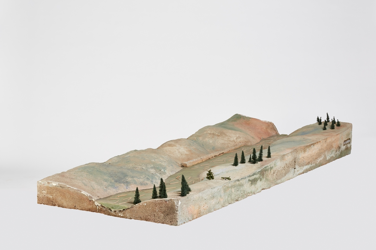 Seksjon to ovenfra (oppstrøms) av modell i fire fysiske seksjoner (FLM.002000).

Modellen som helhet gjengir et elveløp med ulike fløtingsrelaterte bygningselementer (dam, forbygninger, steinkar, lenser, bygninger og broer) og situasjoner (lunnet tømmer, tømmer som fløtes, tømmervaser). Modellen illustrerer ikke et reelt elveløp, men bygningselementene kan være basert på eksisterende sådan. Skala er ikke angitt, men kan beregnes omtrentlig.

Overflaten gjengir en landskapskontur med elvebunn. Elvebunnen er fylt med "vann" i form av et plastmateriale. Landoverflaten er malt i grå og grønne valører for å illustrere berggrunn, løsmasser og vegatasjon. Modelltrær er limt på overflaten. Trepinner er limt på vannflaten for å illustrere tømmerstokker. Det er tre bygninger på modellen - et større bolighus, en mindre hytte eller koie, og en flytende hvilebrakke.

Seksjon en (FLM.002000.A) viser en dam (nåledam) med bilvei over og forbygninger langs elvebredden nedenfor dammen samt et gult bolighus.

Seksjon to (FLM.002000.B) viser laftet, steinfylt forbygninger som avsperrer små viker og et par tømmerlunner.

Seksjon tre (FLM.002000.C) viser en koie med et par robåter på elvebredden, en laftet, steinfylt forbygning som avsperrer en evje og en tømmervase.

Seksjon fire (FLM.002000.D) viser en vei som krysser elven via en fagverksbro, en attholdslense med fem rierkar og en hvilebrakke.