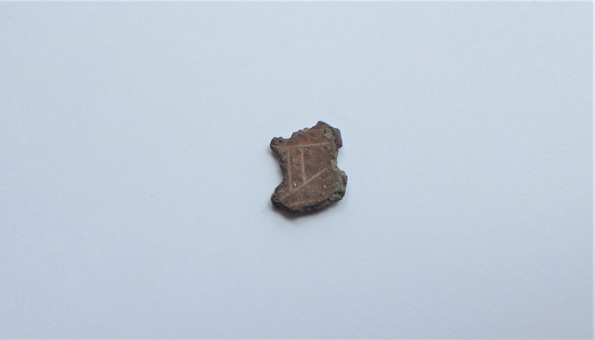 En liten metallplate med bruddflater i alle sider og strekdekor på den ene overflaten.