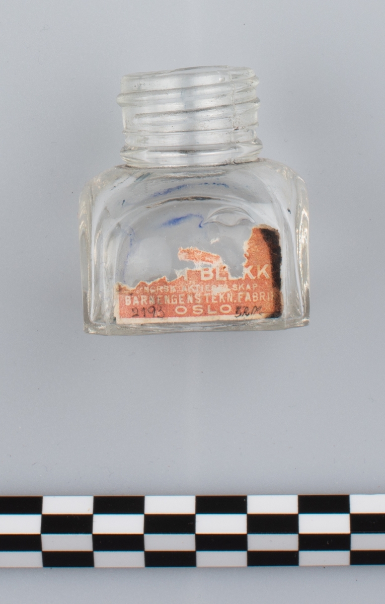 Blank, tom flaske i klart glass uten skrulokk. del av etikett på en side. Rester av blå substans på innsiden. Etiketten har orange bakgrunn