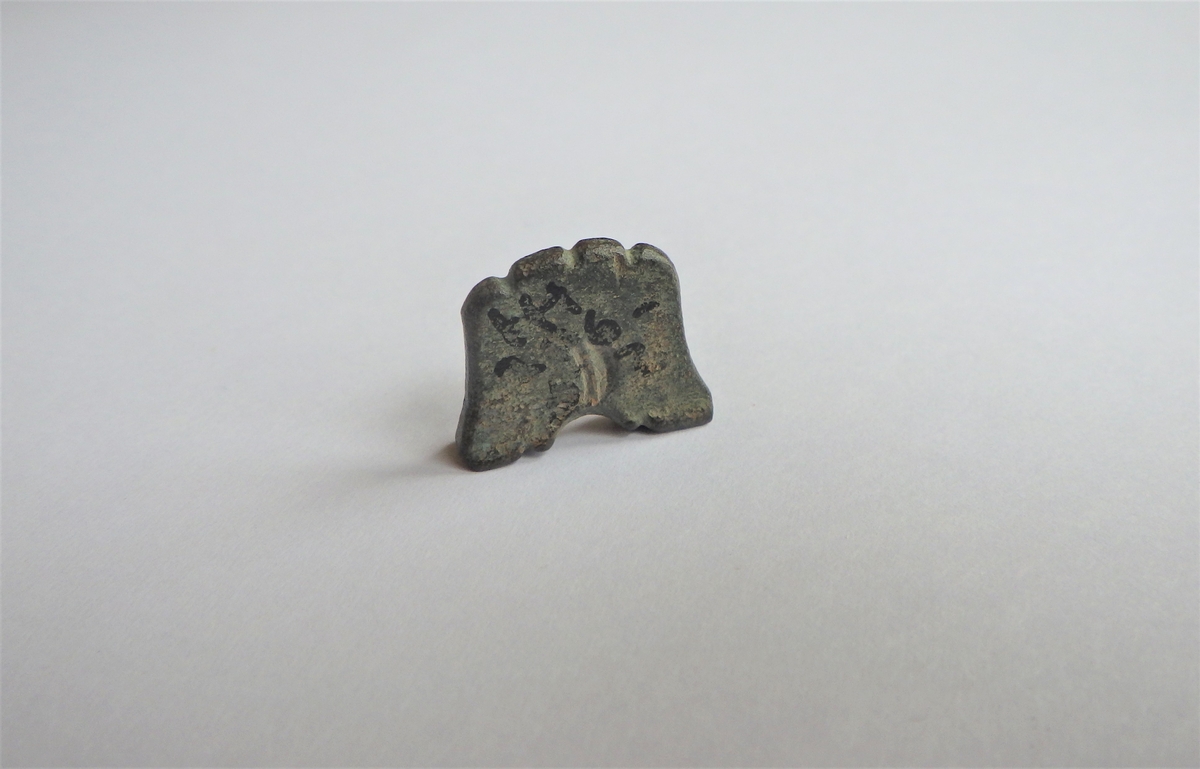 Tilnærmet firkantet gjenstand med en halvsirkelformet fordypning i den tykkeste enden. Denne enden fungerer som en "fot" som gjør at gjenstanden står stabilt oppreist. Tykkelsen er avsmalnende opp fra foten. Alle sider er intakte og toppen, dvs motsatt ende av foten, er bearbeidet med tre fordypninger som gir en bølgeform. Midt i overflaten av gjenstanden, rett over buen, sees på den ene siden rester av en fordypning som kan ha vært et preg. Kan være en spillebrikke.