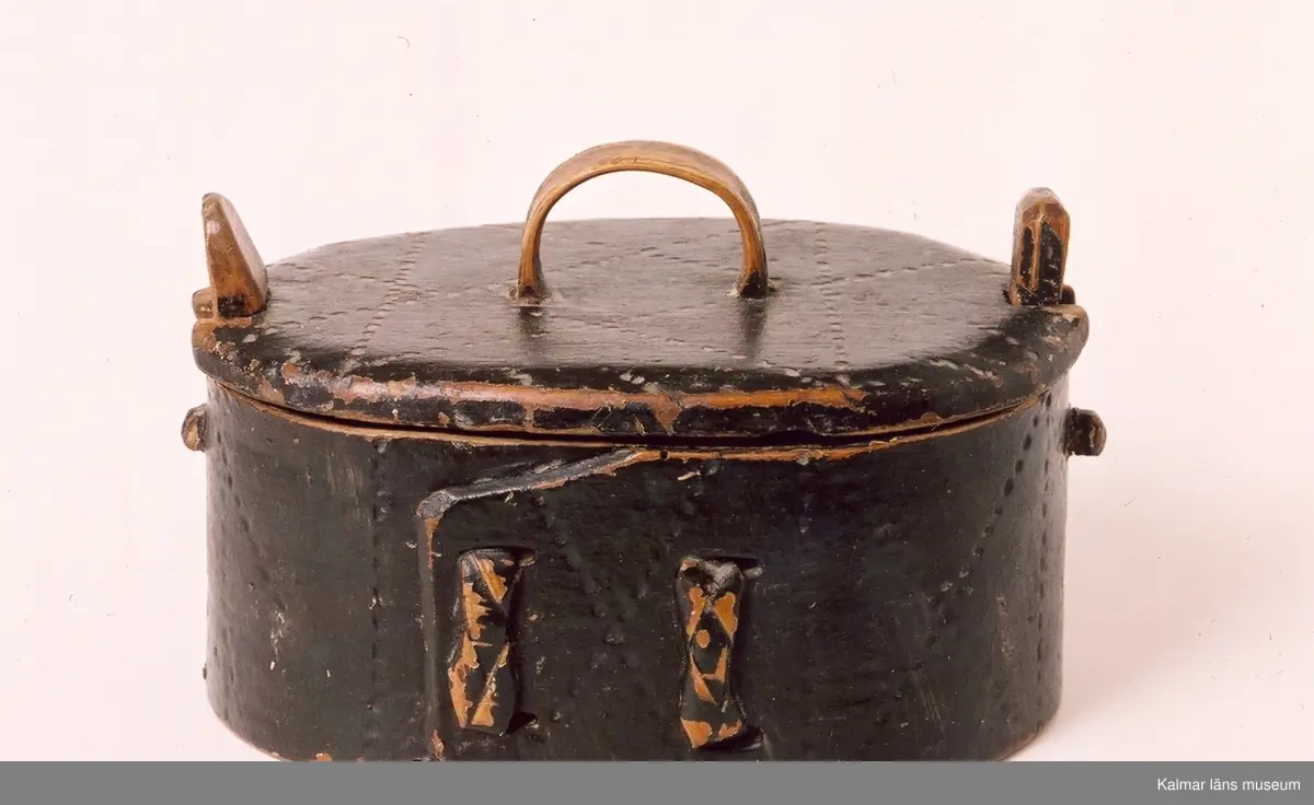 KLM 6754. Äska, svepask med knäpplock och litet handtag på locket. Av böjt bokspån, oval. Ornerad med punktlinjer i geometriskt mönster. Målad helt i mörkgrönt. På lockets insida inskuret 17692 (76 -ihopskrivet), under botten inskuret 1870.