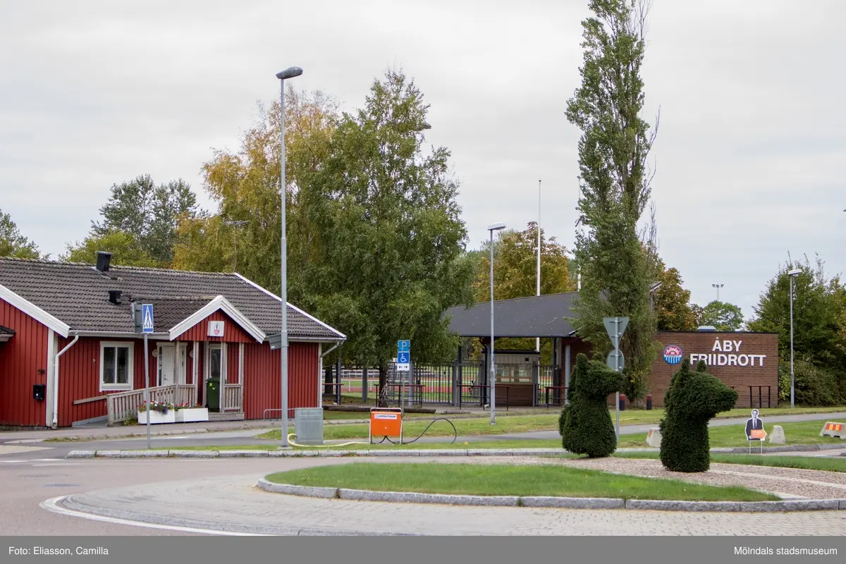 Cirkulationsplats var Idrottsvägen möter Ragnar Thorngrens gata i Åby, Mölndal, den 11 oktober 2016. På andra sidan cirkulationsplatsen ses Mölndals Allmänna Simsällskaps (MASS) klubbhus med adress Idrottsvägen 15 samt idrottsarenan Åby Friidrotts entré med adress Idrottsvägen 13. Vy mot sydväst.