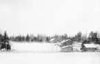 Foto med vy av Strömsdal taget från isen på Sandfjärden i Sankt Anna. Utanför hemmet skymtar figurerna av förre skepparen Anders Österman och makan Ida. Vid tiden för bilden hade Anders sedan en tid slutat med sjömansyrket för att istället driva den handelsbod som syns till höger invid strandkanten nedanför hemmet.