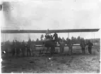 Flygplan Thulin FA ur Flygkompaniet står på ett fält på Malmen, cirka 1918. 13 militärer står vid flygplanet.