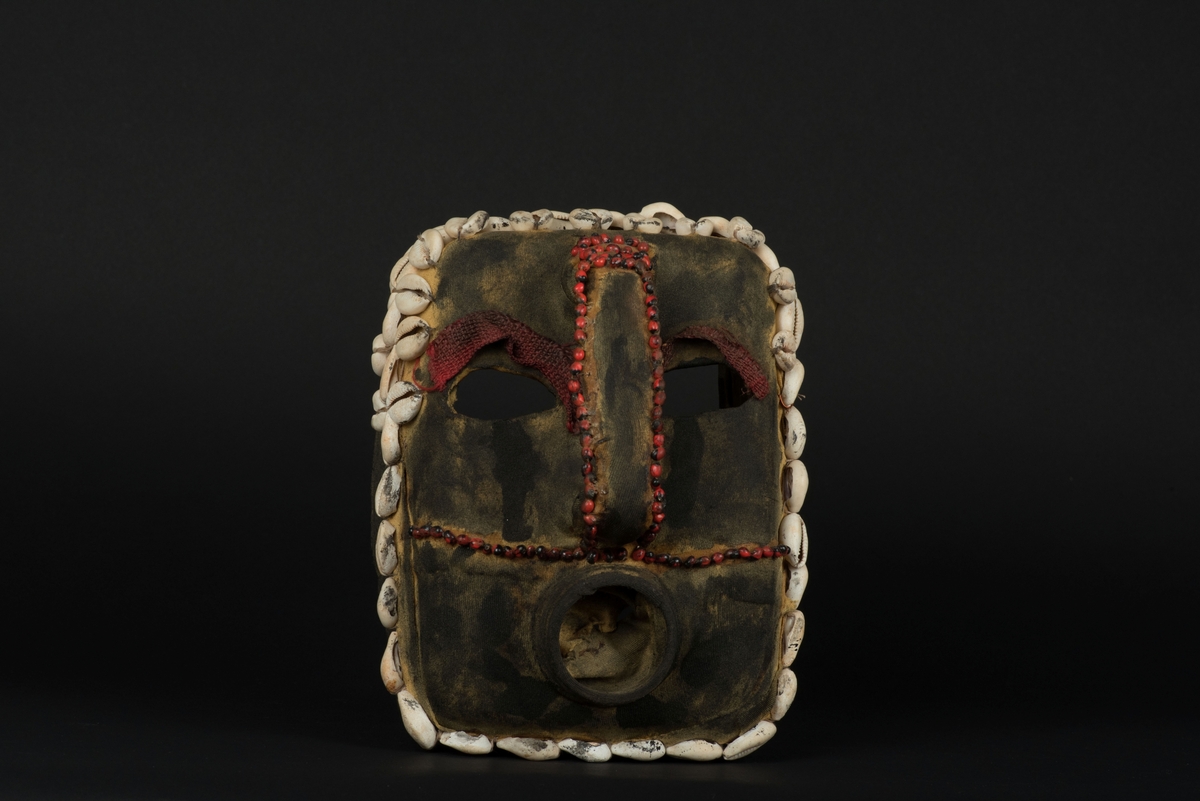 Rektangulär mask tillverkad av en dekorerad överdel av en plastdunk.
Den avskurna dunken är klädd med textil, troligen ett nylontyg, som är delvis svartmålat. Två utskurna hål bildar ögonen på var sida om handtaget som utgör näsan. Dunköppningen därunder blir en mun. Över ögonen sitter pålimmade vävda röda band i plast som ögonbryn. 
Runt ansiktet sitter en rad med kaurissnäckor och runt handtaget/näsan samt på en rad mellan mun och näsa finns fastlimmade röda frön eller bönor.
Snäckorna är fästade med tråd och på något ställe med metalltråd. Bönorna är trädda på ett snöre, sedan limmade.