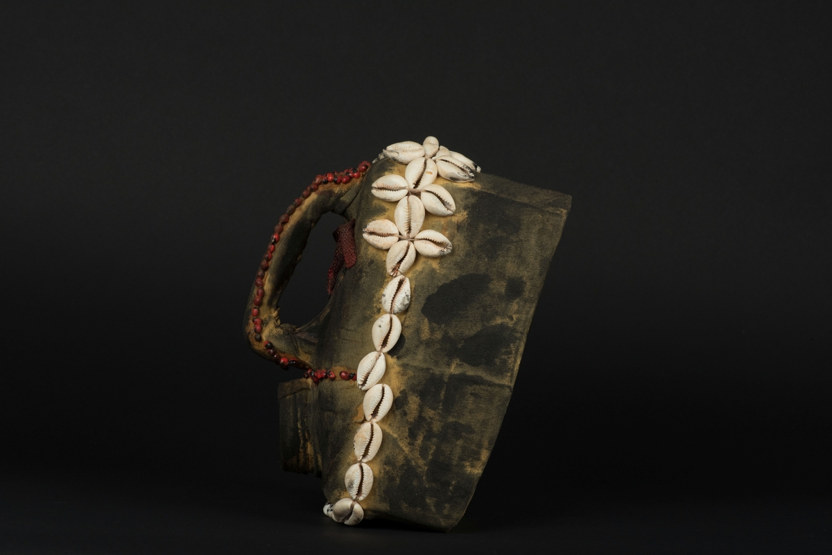 Rektangulär mask tillverkad av en dekorerad överdel av en plastdunk.
Den avskurna dunken är klädd med textil, troligen ett nylontyg, som är delvis svartmålat. Två utskurna hål bildar ögonen på var sida om handtaget som utgör näsan. Dunköppningen därunder blir en mun. Över ögonen sitter pålimmade vävda röda band i plast som ögonbryn. 
Runt ansiktet sitter en rad med kaurissnäckor och runt handtaget/näsan samt på en rad mellan mun och näsa finns fastlimmade röda frön eller bönor.
Snäckorna är fästade med tråd och på något ställe med metalltråd. Bönorna är trädda på ett snöre, sedan limmade.