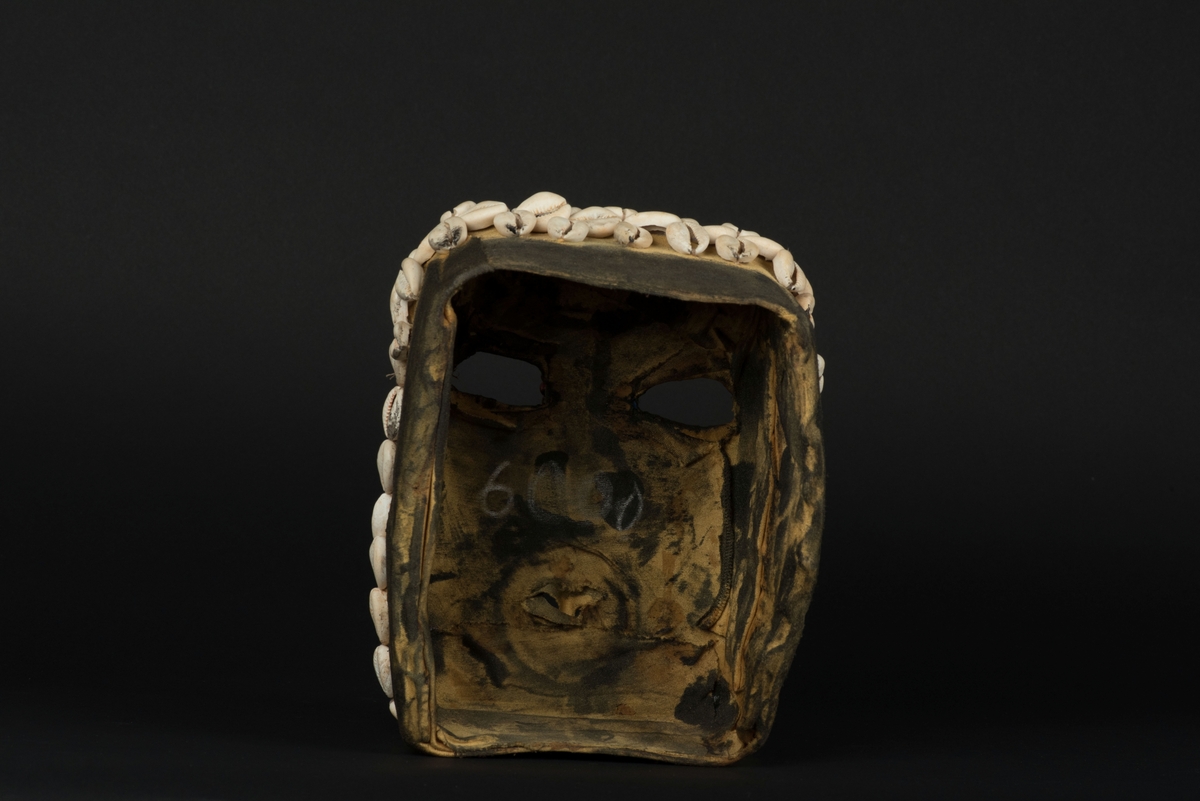 Rektangulär mask tillverkad av en dekorerad överdel av en plastdunk.
Den avskurna dunken är klädd med textil, troligen ett nylontyg, som är delvis svartmålat. Två utskurna hål bildar ögonen på var sida om handtaget som utgör näsan. Dunköppningen därunder blir en mun. Över ögonen sitter pålimmade vävda röda band i plast som ögonbryn. 
Runt ansiktet sitter en rad med kaurissnäckor och runt handtaget/näsan samt på en rad mellan mun och näsa finns fastlimmade röda frön eller bönor.
Snäckorna är fästade med tråd och på något ställe med metalltråd. Bönorna är trädda på ett snöre, sedan limmade.