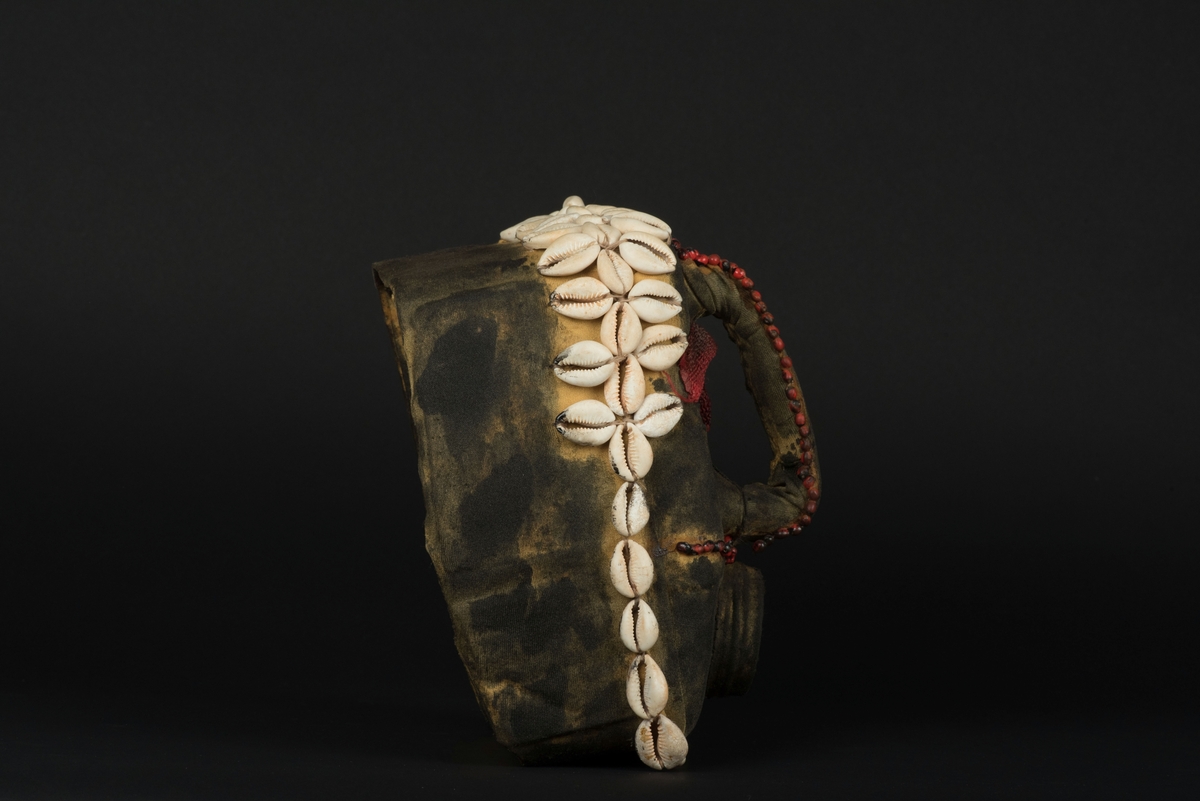 Rektangulär mask tillverkad av en dekorerad överdel av en plastdunk.
Den avskurna dunken är klädd med textil, troligen ett nylontyg, som är delvis svartmålat. Två utskurna hål bildar ögonen på var sida om handtaget som utgör näsan. Dunköppningen därunder blir en mun. Över ögonen sitter pålimmade vävda röda band i plast som ögonbryn. 
Runt ansiktet sitter en rad med kaurissnäckor och runt handtaget/näsan samt på en rad mellan mun och näsa finns fastlimmade röda frön eller bönor.
Snäckorna är fästade med tråd och på något ställe med metalltråd. Bönorna är trädda på ett snöre, sedan limmade.