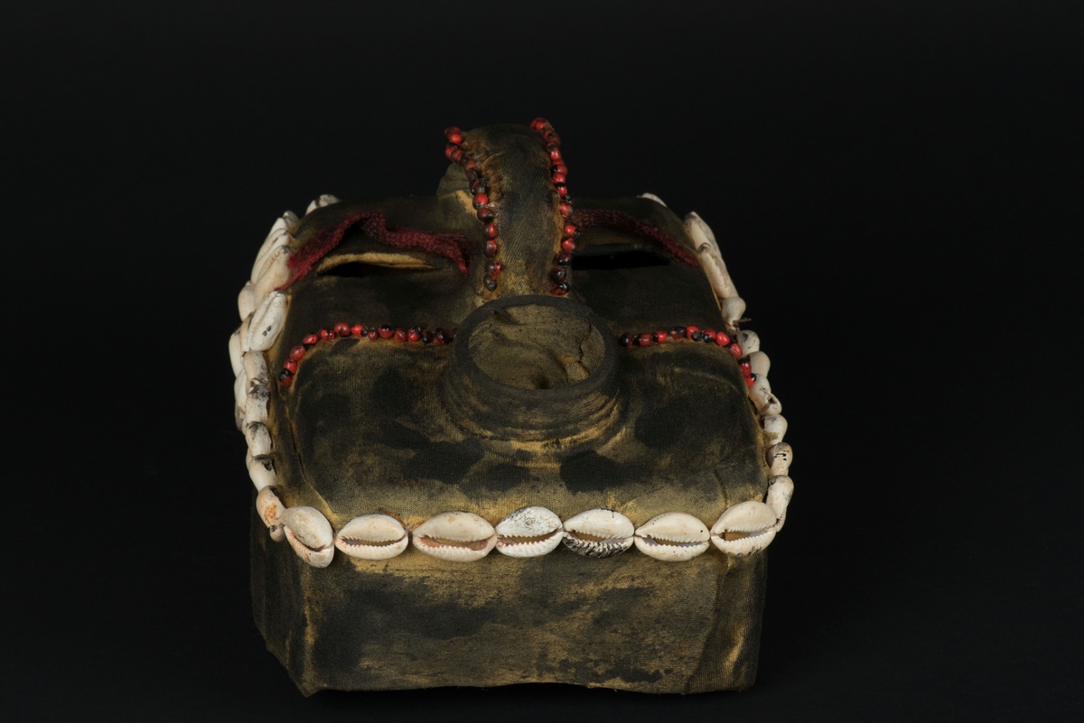 Rektangulär mask tillverkad av en dekorerad överdel av en plastdunk.
Den avskurna dunken är klädd med textil, troligen ett nylontyg, som är delvis svartmålat. Två utskurna hål bildar ögonen på var sida om handtaget som utgör näsan. Dunköppningen därunder blir en mun. Över ögonen sitter pålimmade vävda röda band i plast som ögonbryn. 
Runt ansiktet sitter en rad med kaurissnäckor och runt handtaget/näsan samt på en rad mellan mun och näsa finns fastlimmade röda frön eller bönor.
Snäckorna är fästade med tråd och på något ställe med metalltråd. Bönorna är trädda på ett snöre, sedan limmade.