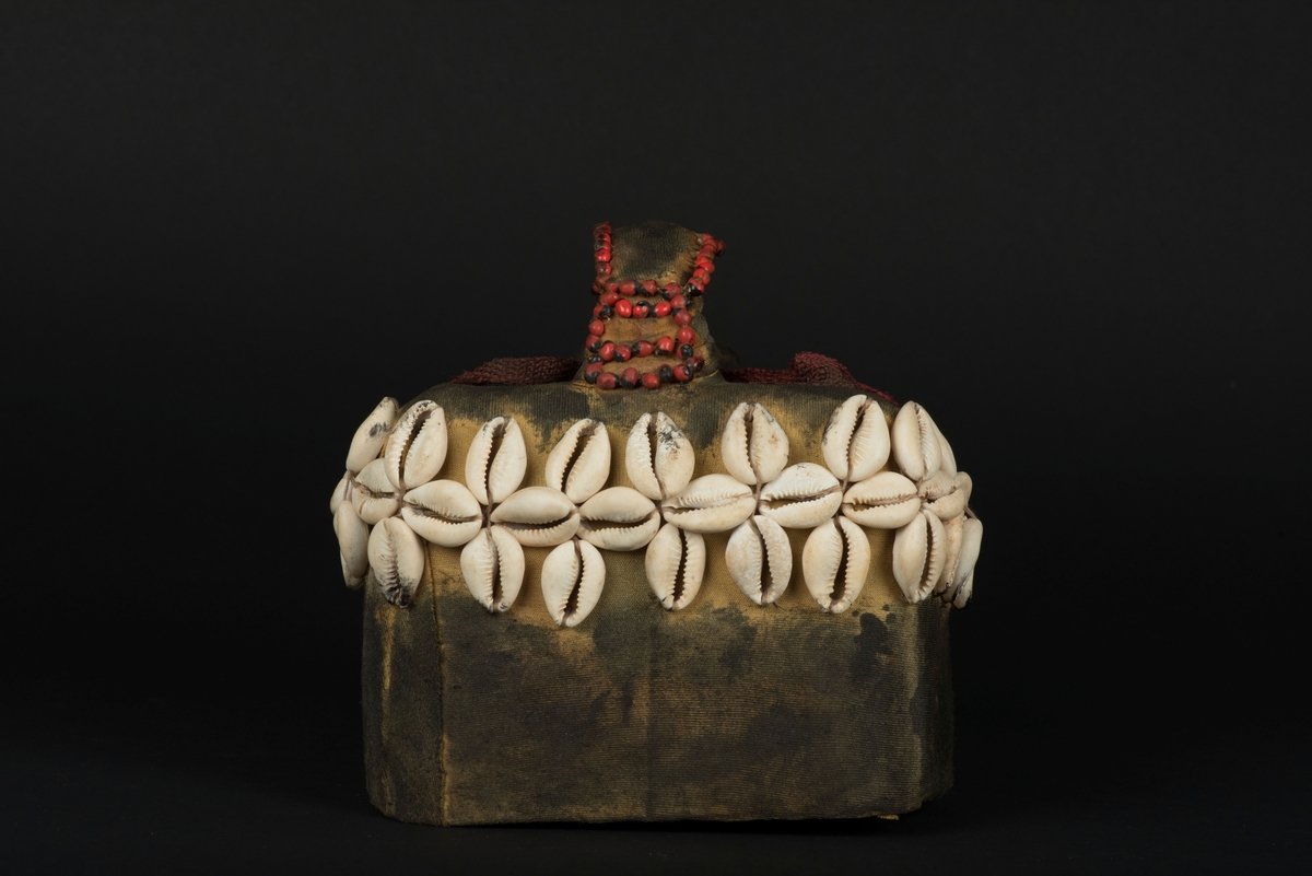 Rektangulär mask tillverkad av en dekorerad överdel av en plastdunk.
Den avskurna dunken är klädd med textil, troligen ett nylontyg, som är delvis svartmålat. Två utskurna hål bildar ögonen på var sida om handtaget som utgör näsan. Dunköppningen därunder blir en mun. Över ögonen sitter pålimmade vävda röda band i plast som ögonbryn. 
Runt ansiktet sitter en rad med kaurissnäckor och runt handtaget/näsan samt på en rad mellan mun och näsa finns fastlimmade röda frön eller bönor.
Snäckorna är fästade med tråd och på något ställe med metalltråd. Bönorna är trädda på ett snöre, sedan limmade.