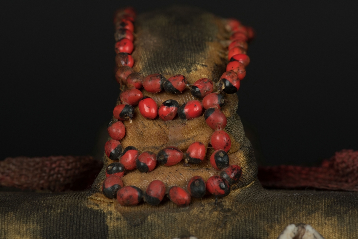 Rektangulär mask tillverkad av en dekorerad överdel av en plastdunk.
Den avskurna dunken är klädd med textil, troligen ett nylontyg, som är delvis svartmålat. Två utskurna hål bildar ögonen på var sida om handtaget som utgör näsan. Dunköppningen därunder blir en mun. Över ögonen sitter pålimmade vävda röda band i plast som ögonbryn. 
Runt ansiktet sitter en rad med kaurissnäckor och runt handtaget/näsan samt på en rad mellan mun och näsa finns fastlimmade röda frön eller bönor.
Snäckorna är fästade med tråd och på något ställe med metalltråd. Bönorna är trädda på ett snöre, sedan limmade.