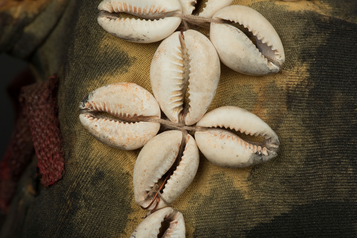 Rektangulär mask tillverkad av en dekorerad överdel av en plastdunk.
Den avskurna dunken är klädd med textil, troligen ett nylontyg, som är delvis svartmålat. Två utskurna hål bildar ögonen på var sida om handtaget som utgör näsan. Dunköppningen därunder blir en mun. Över ögonen sitter pålimmade vävda röda band i plast som ögonbryn. 
Runt ansiktet sitter en rad med kaurissnäckor och runt handtaget/näsan samt på en rad mellan mun och näsa finns fastlimmade röda frön eller bönor.
Snäckorna är fästade med tråd och på något ställe med metalltråd. Bönorna är trädda på ett snöre, sedan limmade.